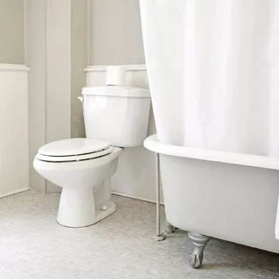 A minimalistic bathroom featuring a white toilet with a roll of toilet paper on top, a white clawfoot bathtub with a partially drawn white shower curtain, light-colored floor tiles, and light-painted walls.